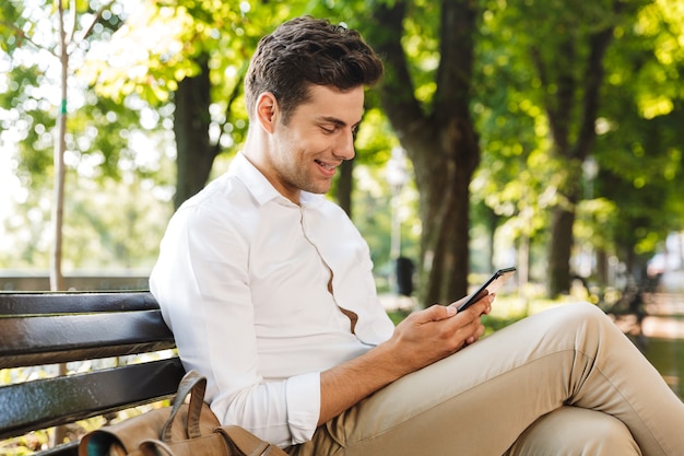Lächelnder junger Geschäftsmann, der draußen auf einer Bank sitzt und Handy betrachtet