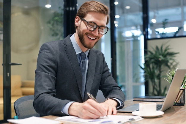 Lächelnder junger Geschäftsmann, der etwas aufschreibt, während er im modernen Büro arbeitet.