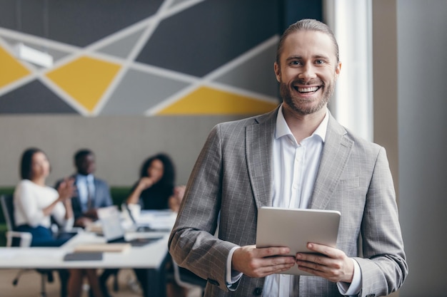 Lächelnder junger Geschäftsmann hält ein digitales Tablet in der Hand, während seine Kollegen im Hintergrund arbeiten