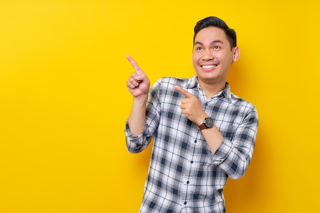 Lächelnder junger gutaussehender asiatischer Mann der 20er Jahre, der ein kariertes Hemd trägt und mit dem Finger auf die Werbefläche zeigt, die auf gelbem Hintergrund isoliert ist Lifestyle-Konzept für Menschen