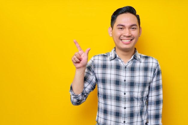 Lächelnder junger, gutaussehender asiatischer Mann, der ein kariertes Hemd trägt und mit dem Finger auf den leeren Kopierbereich zeigt, der auf gelbem Hintergrund isoliert ist Lifestyle-Konzept für Menschen