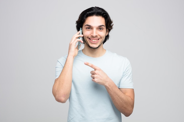 Lächelnder junger gutaussehender Mann, der die Kamera anschaut, die zur Seite zeigt, während er am Telefon spricht, isoliert auf weißem Hintergrund