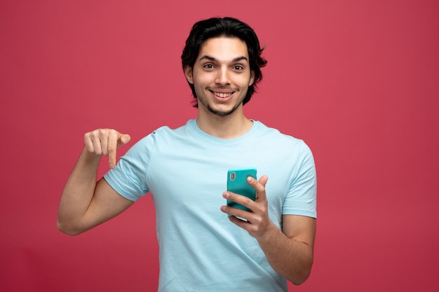 Lächelnder junger gutaussehender Mann mit Handy, der auf die Kamera blickt, die nach unten zeigt, isoliert auf rotem Hintergrund