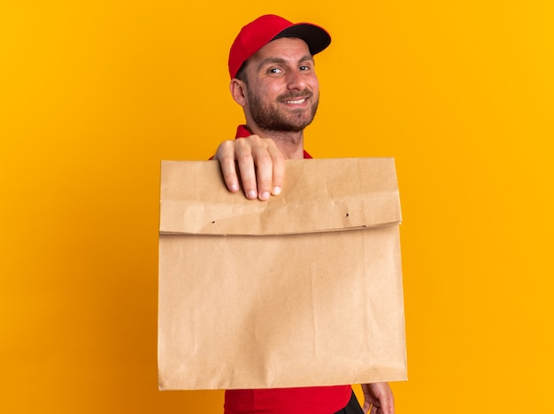 Lächelnder junger kaukasischer Lieferbote in roter Uniform und Mütze, der in der Profilansicht steht und auf die Kamera schaut, die das Papierpaket in Richtung der Kamera ausstreckt, die auf der orangefarbenen Wand isoliert ist?