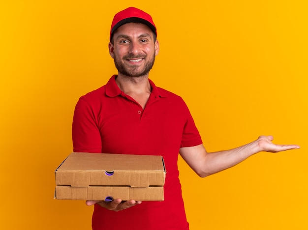 Lächelnder junger kaukasischer Lieferbote in roter Uniform und Mütze mit Blick auf die Kamera, die Pizzapakete hält, die leere Hand isoliert auf oranger Wand zeigen?