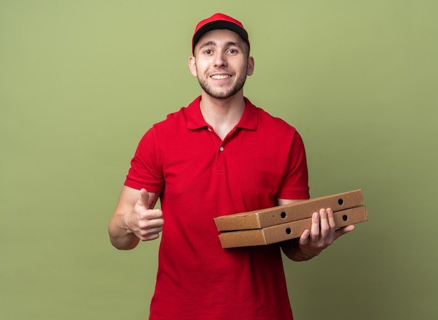 Lächelnder junger Lieferbote in Uniform mit Mütze mit Pizza, die Daumen nach oben zeigt
