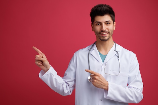 Lächelnder junger männlicher Arzt mit medizinischer Uniform und Stethoskop um den Hals, der auf die Kamera zeigt, die auf die Seite zeigt, die auf rotem Hintergrund isoliert ist