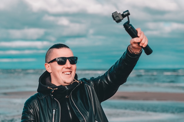 Lächelnder junger männlicher Blogger in Sonnenbrillen, die Selfie machen oder Video am Strand mit Aktionskamera mit kardanischem Kamerastabilisator streamen.