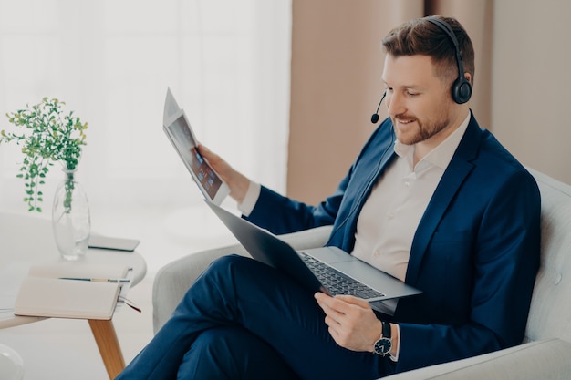 Lächelnder junger männlicher Chef im Anzug mit Headset, um Videoanrufe mit Mitarbeitern zu tätigen, auf den Laptop-Bildschirm zu schauen und den Bericht zu diskutieren, während er im Sessel im Wohnzimmer seiner Wohnung sitzt