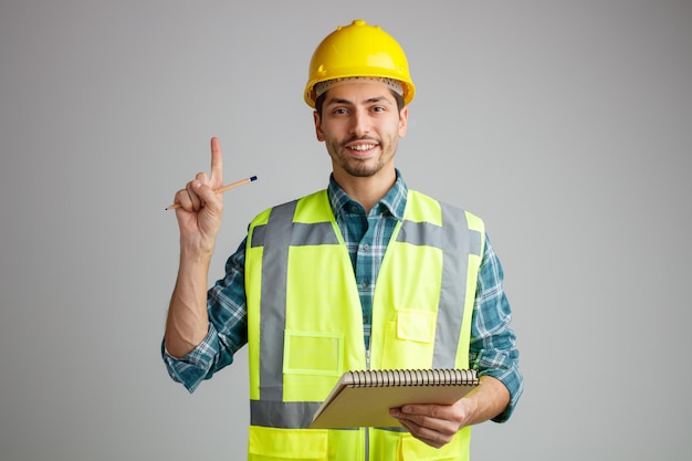 Lächelnder junger männlicher Ingenieur mit Schutzhelm und Uniform, der in die Kamera blickt und Notizblock und Bleistift nach oben zeigt, isoliert auf weißem Hintergrund