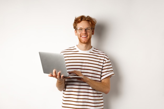 Lächelnder junger Mann, der an Laptop arbeitet und freudig schaut, über weißem Hintergrund stehend.