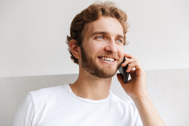 Lächelnder junger Mann, der auf Handy spricht und zu Hause an der Wand steht