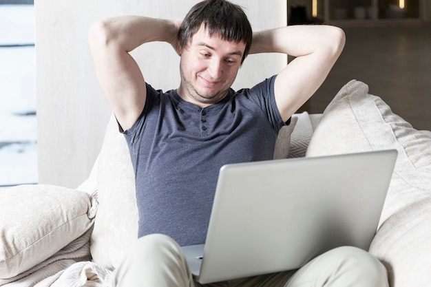 Lächelnder junger Mann, der auf Sofa mit Laptop sitzt. Fernarbeit während der Coronavirus-Pandemie.