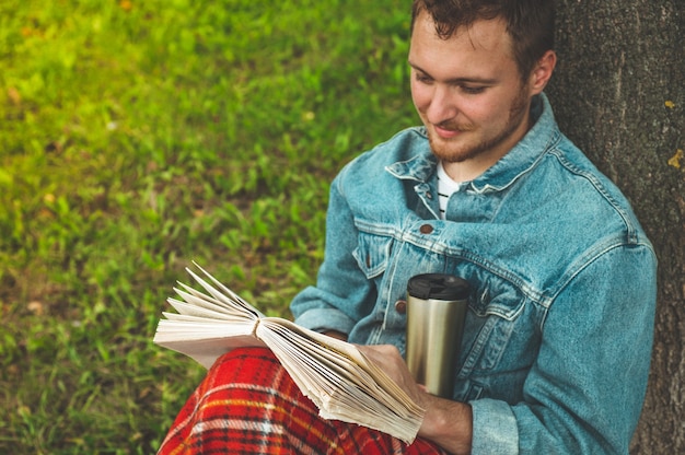 Lächelnder junger Mann, der Buch draußen mit einem warmen roten Plaid und einer Tasse Tee liest. Herbstferien und Lifestyle-Konzept.