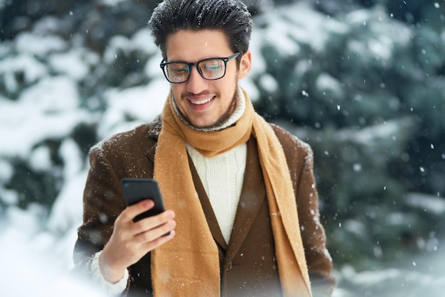 Lächelnder junger Mann, der draußen Spaß hat. Geschäftsmann benutzt ein Telefon. Winterkonzept. Schneefall.