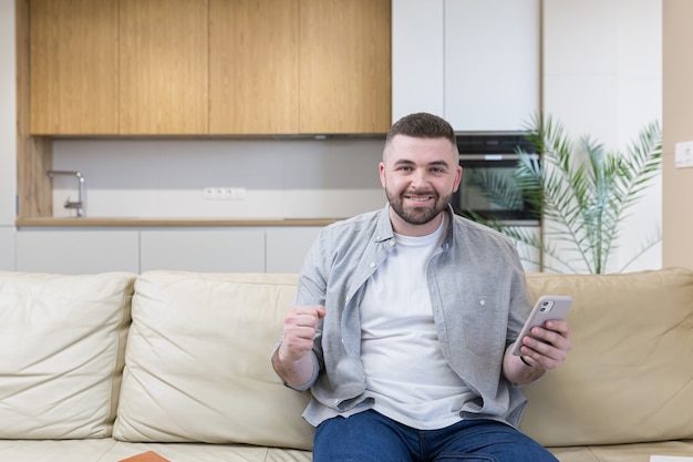Lächelnder junger Mann, der Handy hält, während er auf einem Sofa zu Hause sitzt und feiert