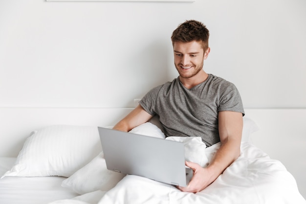 Lächelnder junger Mann, der Laptop beim Sitzen im Bett verwendet