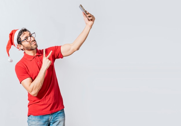 Lächelnder junger Mann in Weihnachtsmütze, der ein selfie lokalisiert nimmt Glücklicher gutaussehender Mann in Weihnachtsmütze, der ein selfie nimmt Glücklicher Kerl, der ein Weihnachtsselfie auf lokalisiertem Hintergrund nimmt