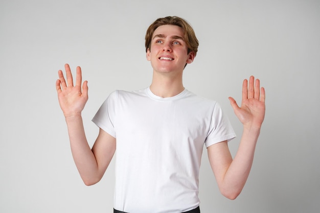 Lächelnder junger Mann in weißem T-Shirt, der mit erhobenen Händen gegen einen einfachen Hintergrund gestimmt
