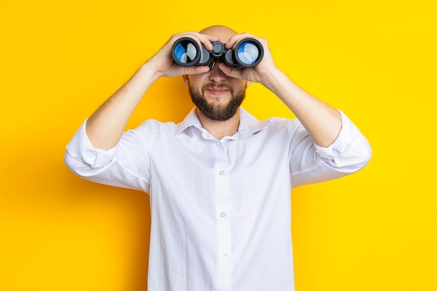 Lächelnder junger Mann mit Bart, der durch ein Fernglas auf gelbem Hintergrund schaut