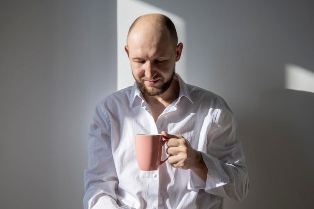 Lächelnder junger Mann mit Bart schaut in eine Tasse Kaffee auf dem Hintergrund einer weißen Wand