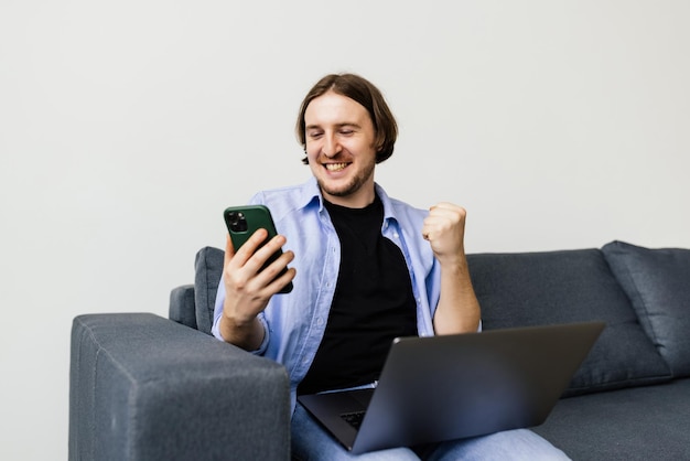 Lächelnder junger Mann mit Handy, während er zu Hause mit Laptop auf einem Sofa sitzt und feiert