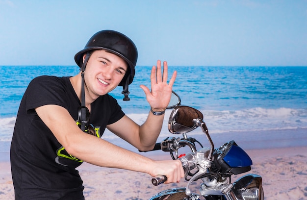 Lächelnder junger Mann mit Helm sitzt auf dem Motorrad am Strand und winkt in die Kamera