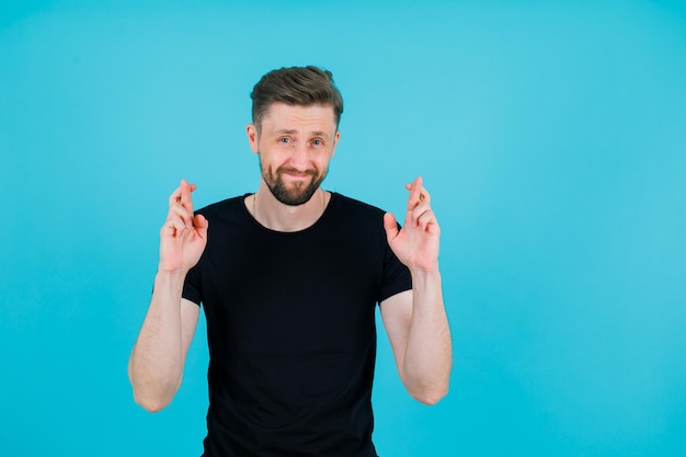 Lächelnder junger Mann schaut in die Kamera, indem er die Finger auf blauem Hintergrund kreuzt