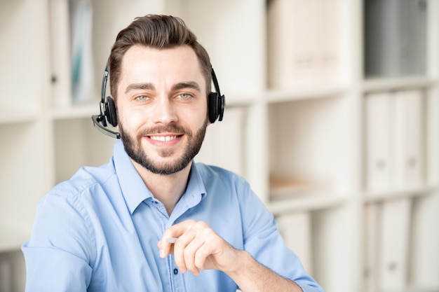 Lächelnder junger Operator mit Kopfhörer, der Sie beim Arbeiten mit Kundenfragen und -anfragen im Büro ansieht