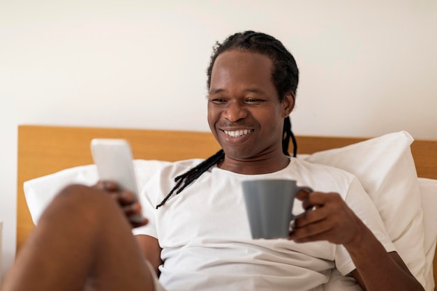 Lächelnder junger schwarzer Mann, der mit Smartphone und Kaffee im Bett ruht