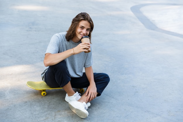 Lächelnder junger Teenager, der Zeit im Skatepark verbringt, auf einem Skateboard sitzt und Kaffee zum Mitnehmen trinkt