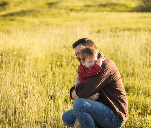 Lächelnder junger Vater und Sohn umarmen sich entspannend auf der Wiese. glücklicher Vater und kleiner Sohn ruhen auf dem Feld des Federgrases