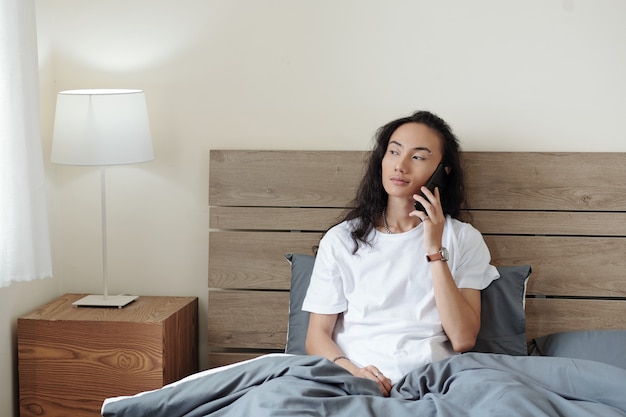 Lächelnder junger vietnamesischer Mann im weißen T-Shirt, der auf dem Bett im Hotelzimmer sitzt und telefonisch anruft