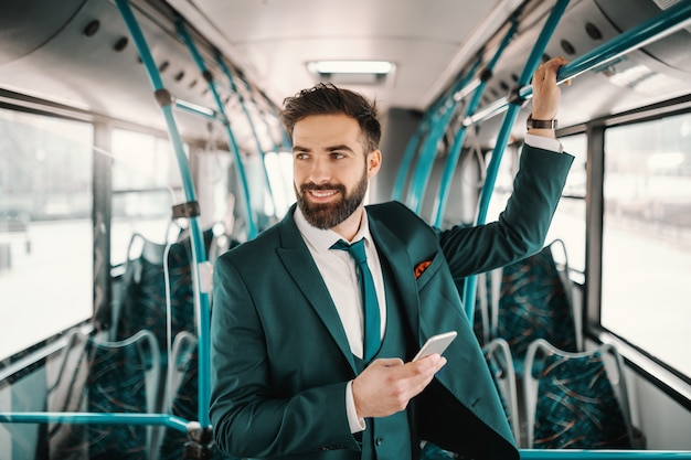 Lächelnder kaukasischer Geschäftsmann im Anzug, der im öffentlichen Bus fährt und Smartphone verwendet. Eines Tages wird es Sinn machen.