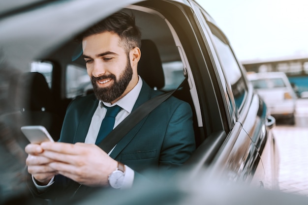 Lächelnder kaukasischer Geschäftsmann in der formellen Abnutzung unter Verwendung des Smartphones beim Sitzen in seinem Auto auf dem Parkplatz.