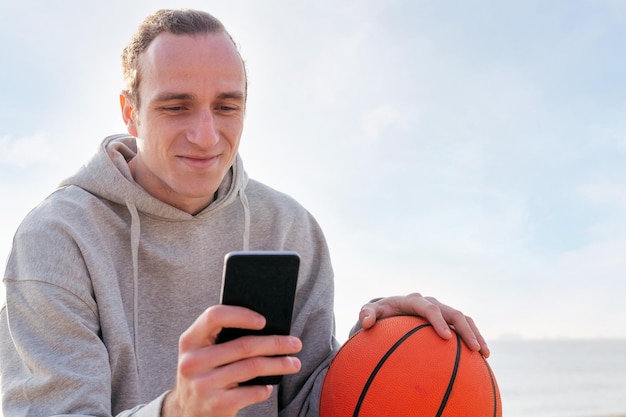 Lächelnder kaukasischer Mann mit Hoodie und Basketball, der sein Handy-Konzept von Lifestyle und Kommunikationstechnologie verwendet, kopieren Sie Platz für Text