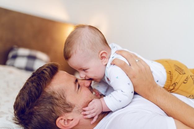 Lächelnder kaukasischer Vater, der auf Bett im Schlafzimmer liegt und seinen liebenden entzückenden Sohn auf seiner Brust hält.