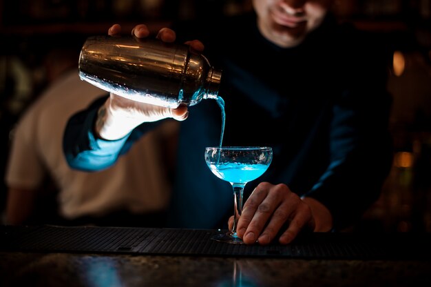 Lächelnder Kellner, der neues Getränk mit blauer Flüssigkeit von einem Schüttel-Apparat in ein Glas unter Verwendung des Siebs gießt