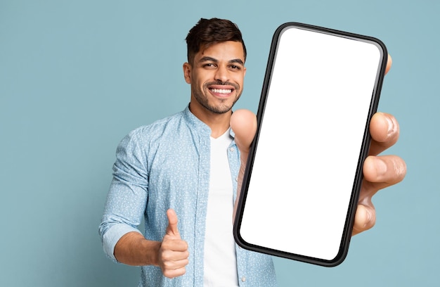 Lächelnder Kerl, der Smartphone mit dem Daumen des leeren Bildschirms oben zeigt