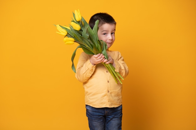 Lächelnder kleiner Junge auf gelbem Studiohintergrund. Fröhliches glückliches Kind mit Tulpenblumenstrauß.