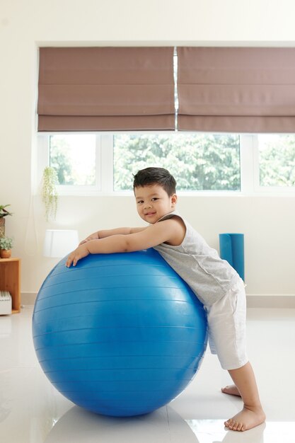 Lächelnder kleiner Junge, der zu Hause neben einem großen Fitnessball steht und in die Kamera schaut