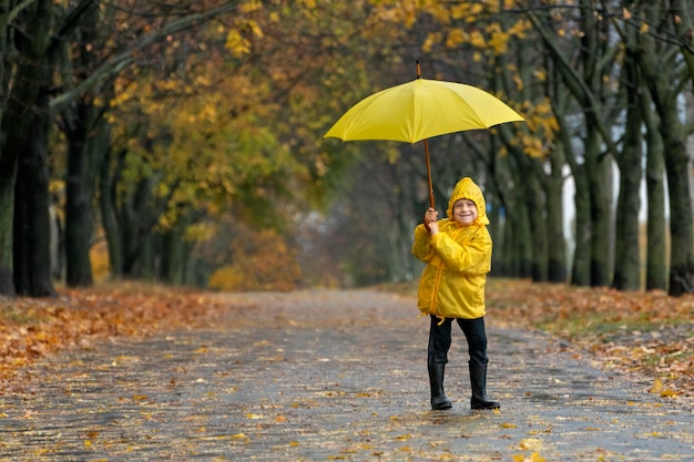 Lächelnder kleiner Junge in einem gelben Regenmantel, der mit einem Regenschirm im Park spazieren geht. Herbstlicher regnerischer Tag