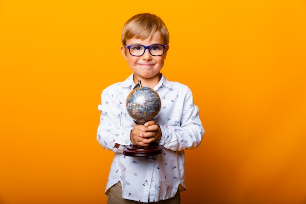 Lächelnder kleiner Junge in Gläsern, der einen Globus auf gelbem Hintergrund hält, Studioporträt eines Kindes.
