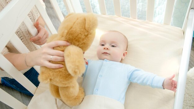 Lächelnder kleiner Junge mit blauen Augen hält und spielt mit Teddybär in der Wiege. Konzept der Erziehung von Familienglück und Babyentwicklung