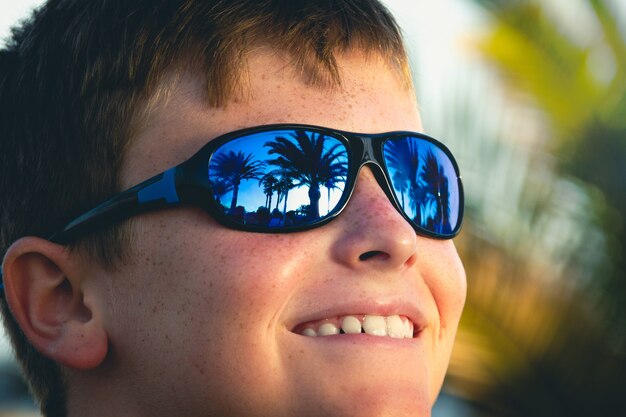 Lächelnder kleiner Junge mit blauer Sonnenbrille am sonnigen Tag im Freien Palmenreflexion auf Brillen