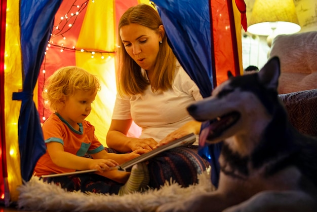 Lächelnder kleiner Junge mit Mutter liest zu Hause im Zelt mit Girlanden ein Buch