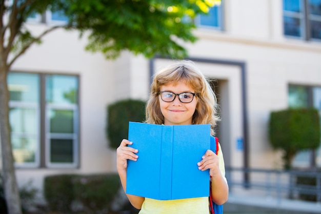 Lächelnder kleiner Studentenjunge, der einen Schulrucksack trägt und ein Schulbuchporträt eines glücklichen Schülers hält