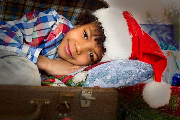 Lächelnder kleiner Weihnachtsmann neben Geschenken. Weihnachtskind, das nahe Geschenken legt. Glückslieferung zu Ihren Diensten. Magische Urlaubsmomente.