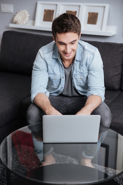 Lächelnder lässiger Mann, der am Laptop-Computer arbeitet, während er zu Hause auf dem Sofa sitzt