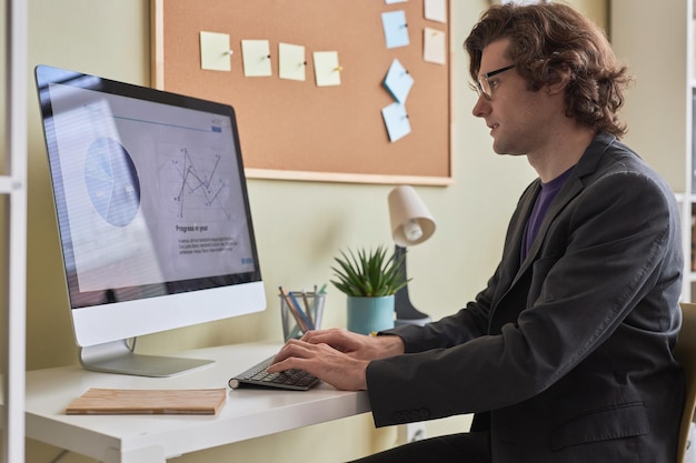 Lächelnder langhaariger Mann benutzt Computer am Schreibtisch im Büro oder zu Hause am Arbeitsplatz